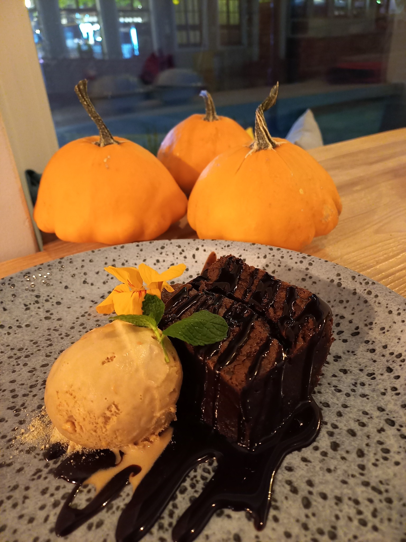 Porcja brownie polanego czekoladą, z gałką lodów, dekoracją z kwiatów i listkiem mięty na talerzu w tle trzech małych dyń.