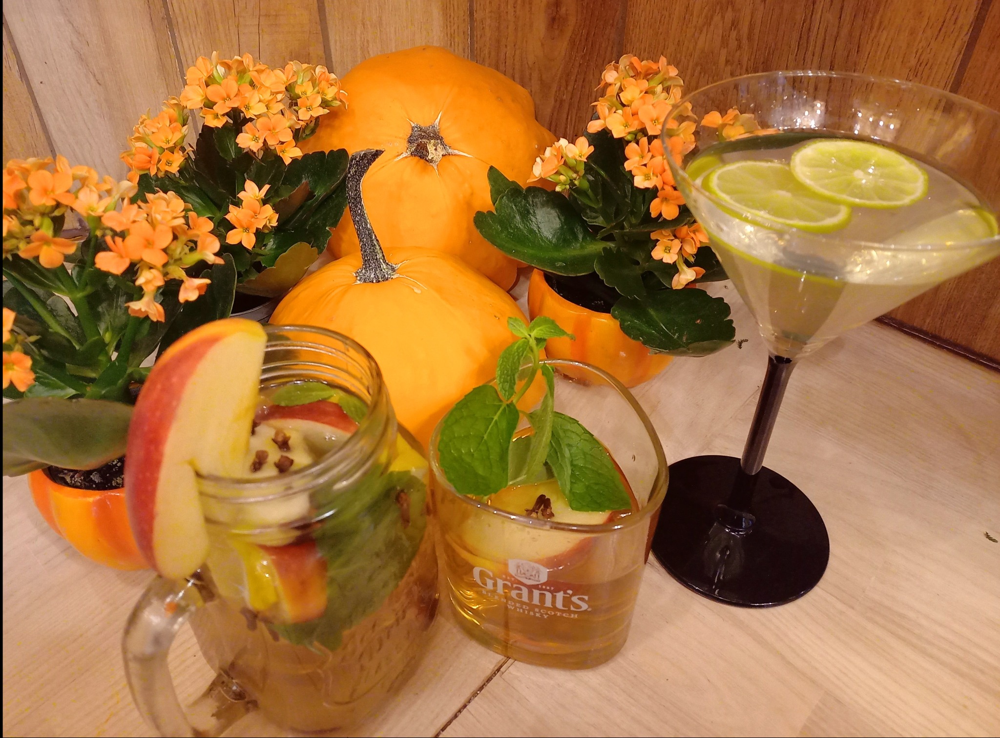 Jesienna kompozycja w Olsztynie: dynie, pomarańczowe kwiaty w doniczkach, koktajle z jabłkami, miętą i limonką w szklankach, jeden z plasterkiem jabłka na krawędzi, drugi w kieliszku do martini.