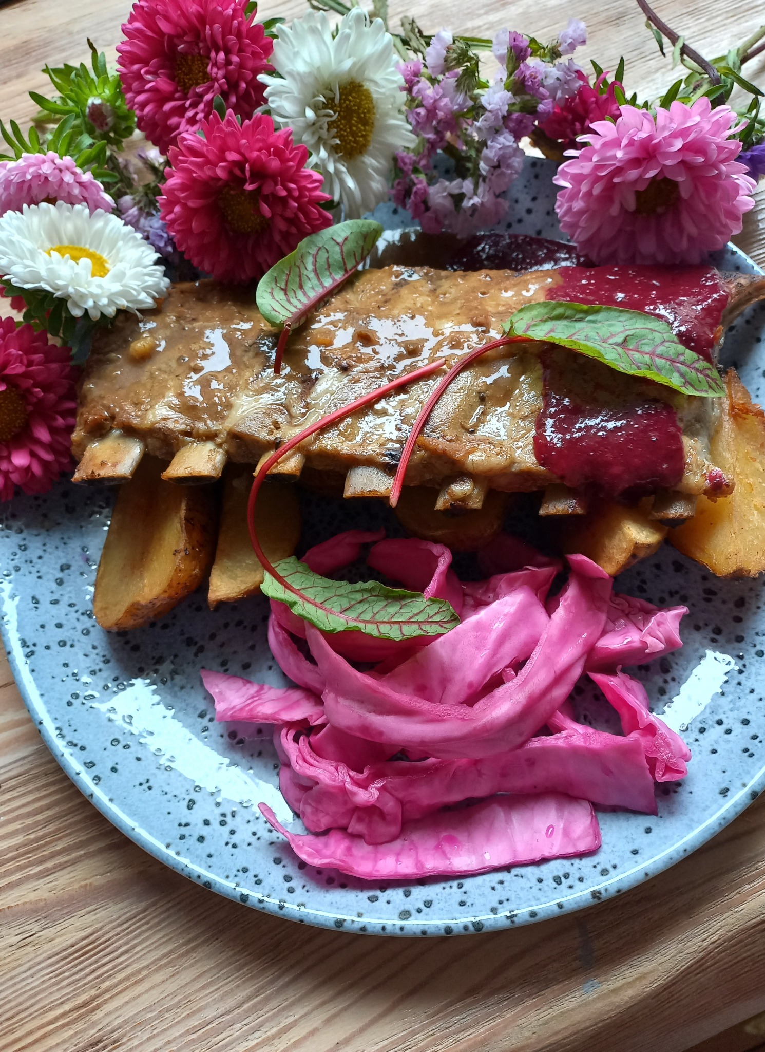 Żeberka w sosie z pieczonymi ziemniakami i różową kapustą na talerzu w kwiatowej oprawie.