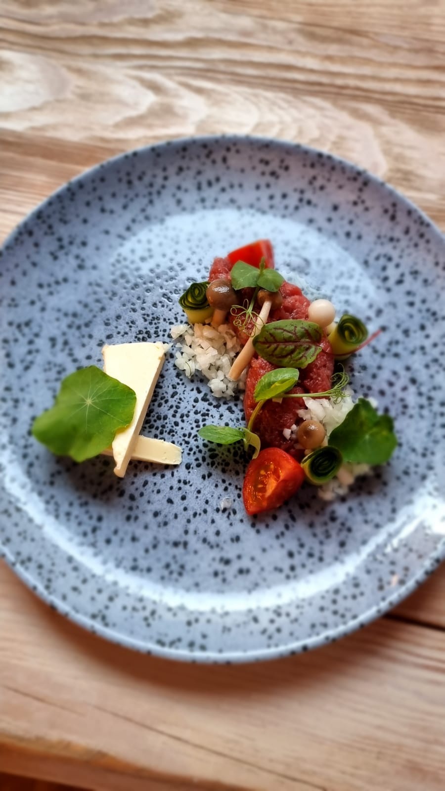 Wykwintnie zaaranżowany tatar wołowy z dodatkami: marynowane grzyby, pomidory cherry, plasterki sera i dekoracyjne liście na talerzu w odcieniach szarości z drobnymi plamkami.