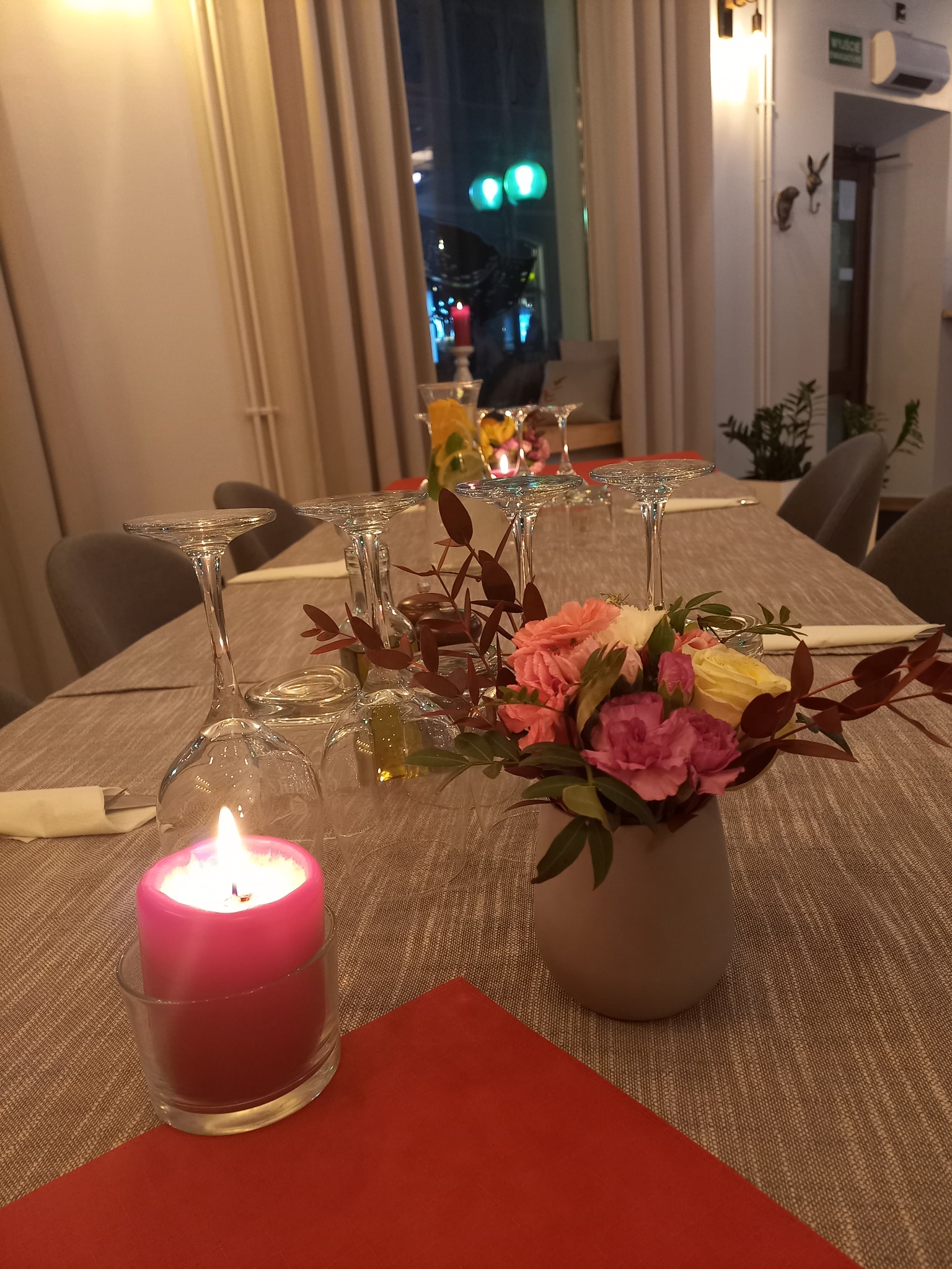 Ustawiony stół z kieliszkami do wina, bukietem kwiatów w szarym wazonie i płonącą różową świecą w szklanym świeczniku, na tle okna z nocnym widokiem.