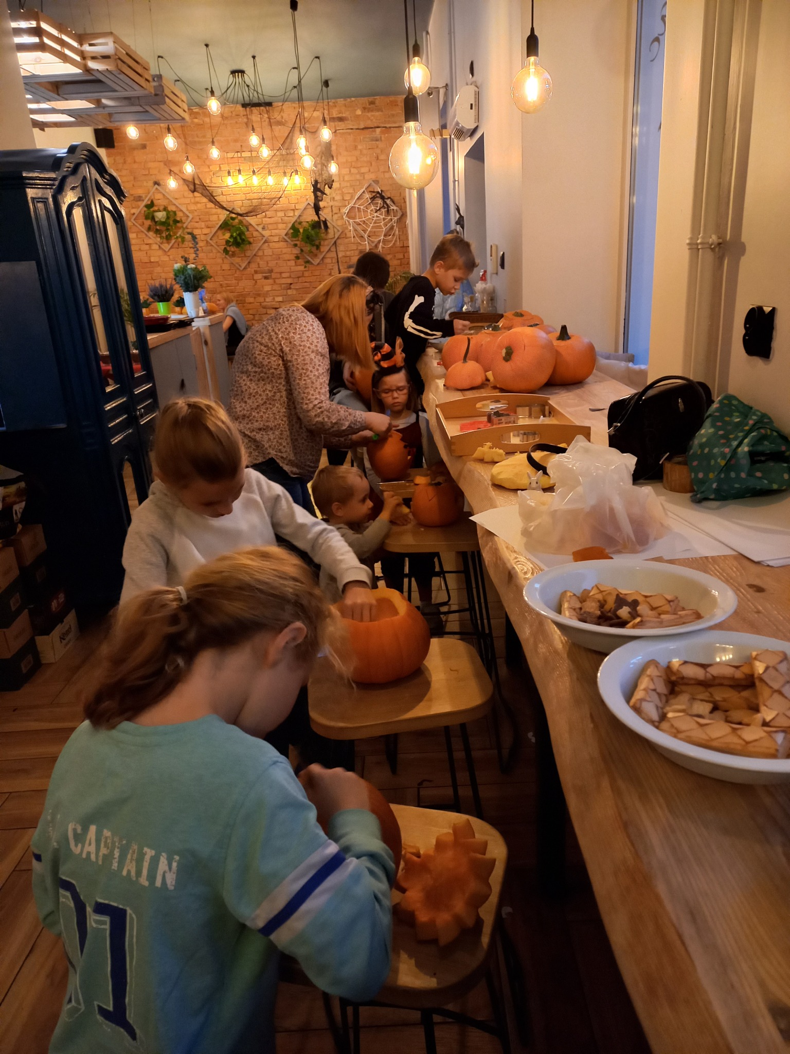 Grupa dzieci i dorosłych wspólnie wydrążają dynie na Halloween przy drewnianym stole, wnętrze z ceglaną ścianą i dekoracyjnym oświetleniem.