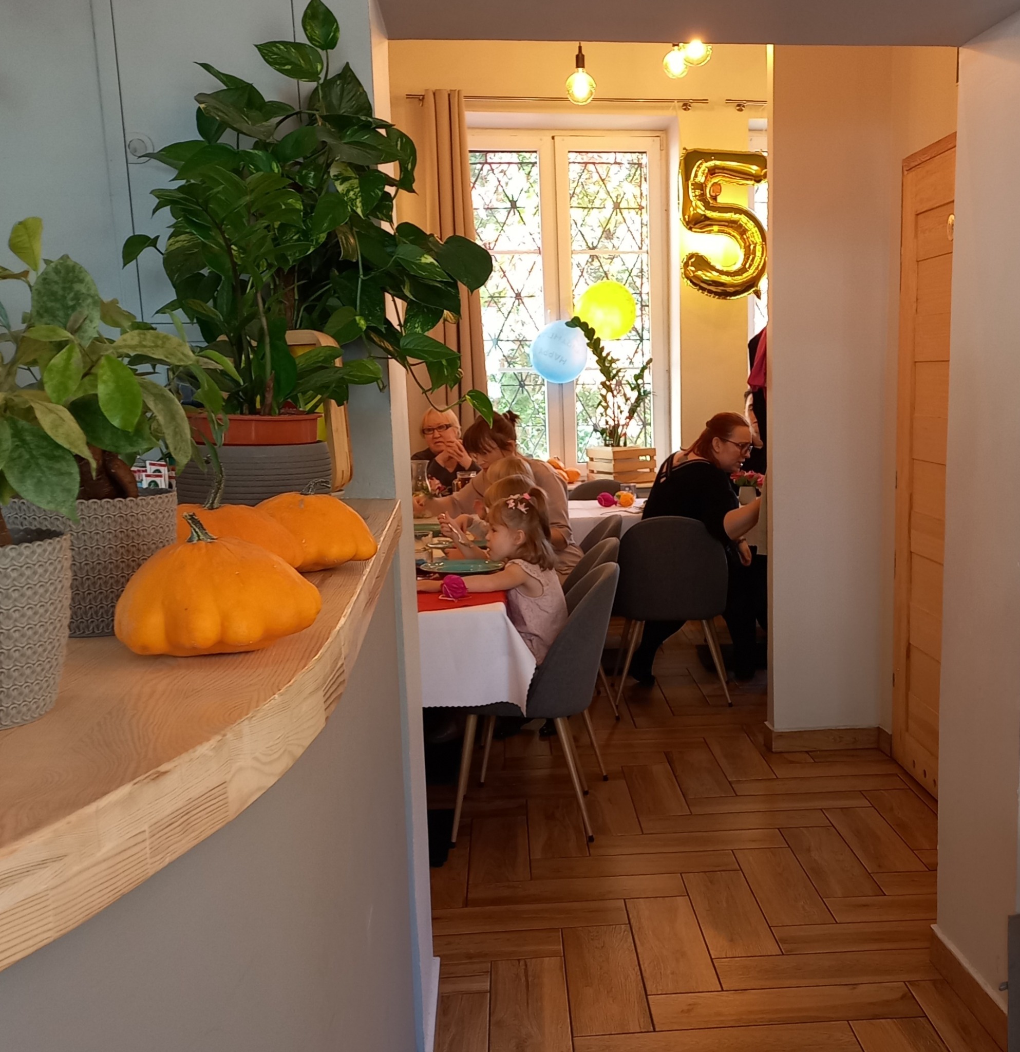Uroczystość urodzinowa w domu: stół nakryty białym obrusem, dzieci siedzące przy stole, dekoracje balonowe, złoty balon w kształcie cyfry 5, dynie ozdobne na półce, rośliny doniczkowe.