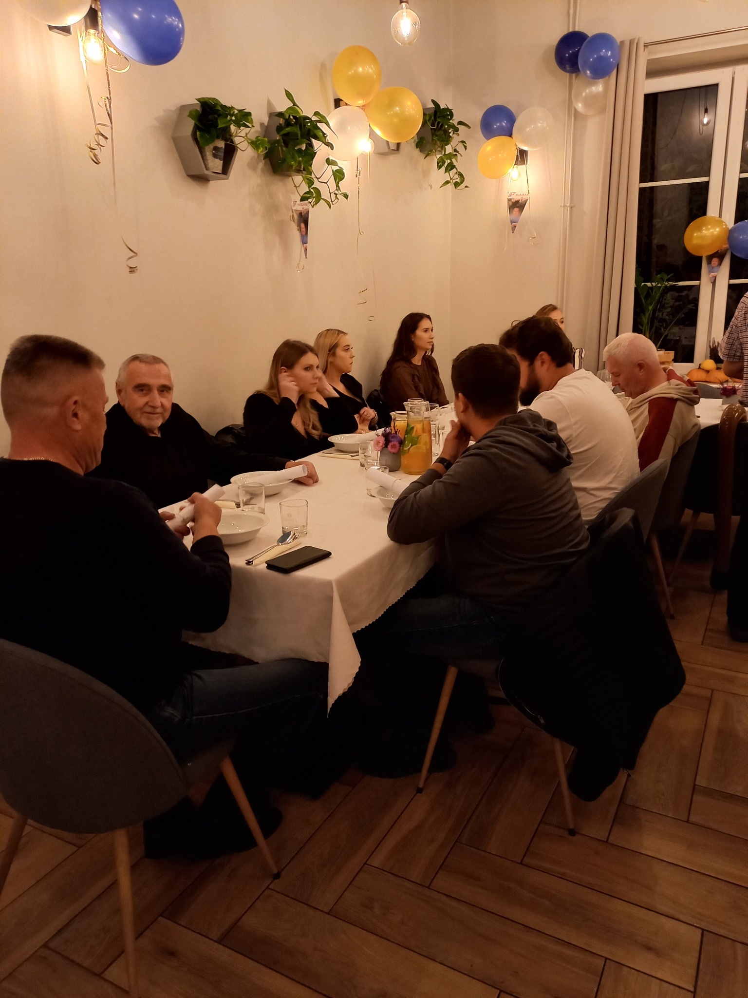 Uczestnicy siedzą przy stole nakrytym białym obrusem, dekorowanym balonami i roślinami w doniczkach, podczas uroczystości.