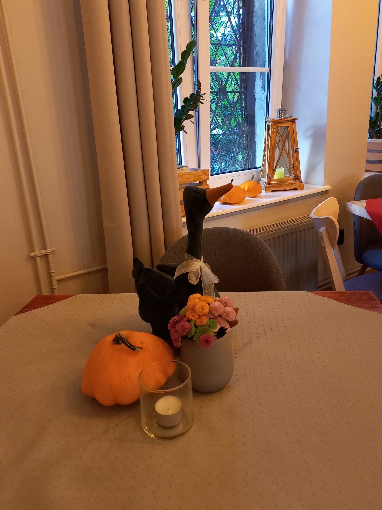 Dekoracja stołu z czarną gęsią z kokardą, dynią, kwiatami w szarym wazonie i świecą w szklanym świeczniku, w tle okno z drewnianą latarenką i dyniami.