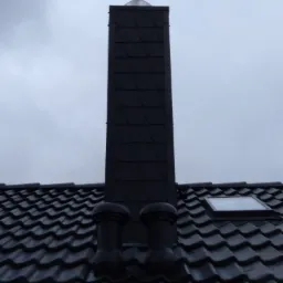 Ciemny dach z dachówki ceramicznej z dwoma kominkami wentylacyjnymi i wysokim kominem z blachy łuskowej, widok z dołu w pochmurny dzień.