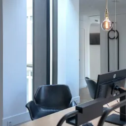 Nowoczesne biuro z drewnianym biurkiem, czarnymi krzesłami i wiszącymi lampami z żarówkami Edisona; na ścianie plakat z Gdynią.