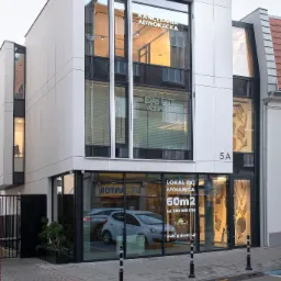 Nowoczesny budynek z dużymi oknami, na których widnieje napis 'Kancelaria Adwokacka', a także informacja o lokalu do wynajęcia, z zaparkowanym samochodem w tle.