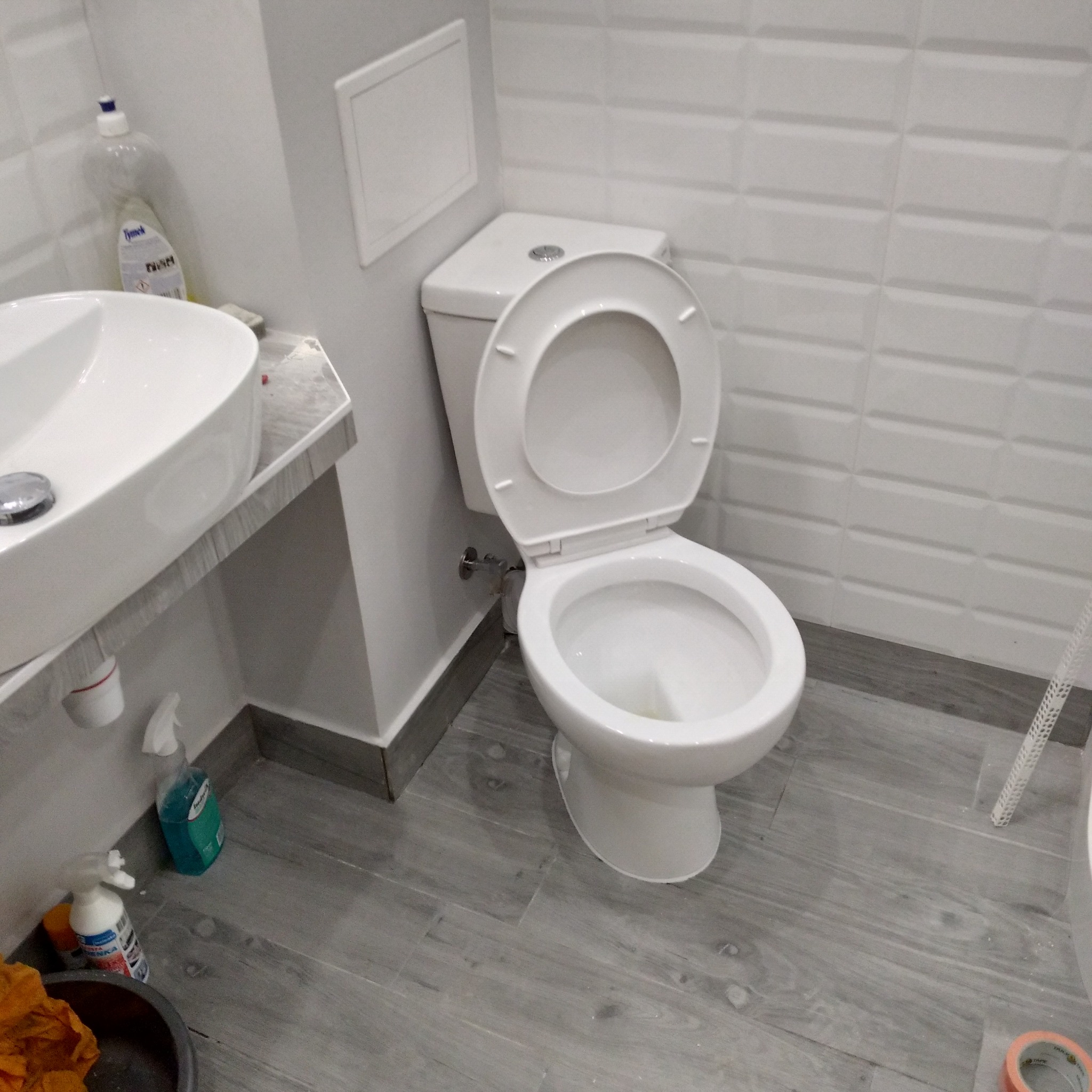 Biała toaleta z otwartą klapą, umywalka i butelka płynu do czyszczenia w odnowionej łazience z szarymi panelami podłogowymi i kafelkami na ścianach.