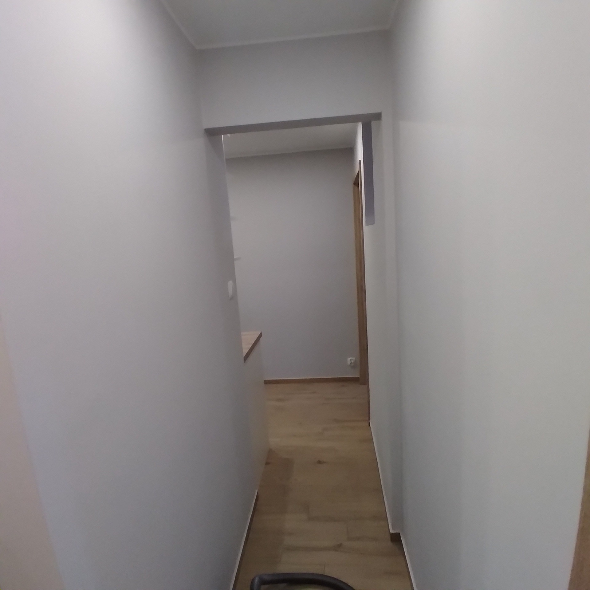 Korytarz po remoncie z gładkimi, szarymi ścianami, drewnianą podłogą i futryną drzwiową bez drzwi. Widoczne gniazdko elektryczne i fragment blatu.