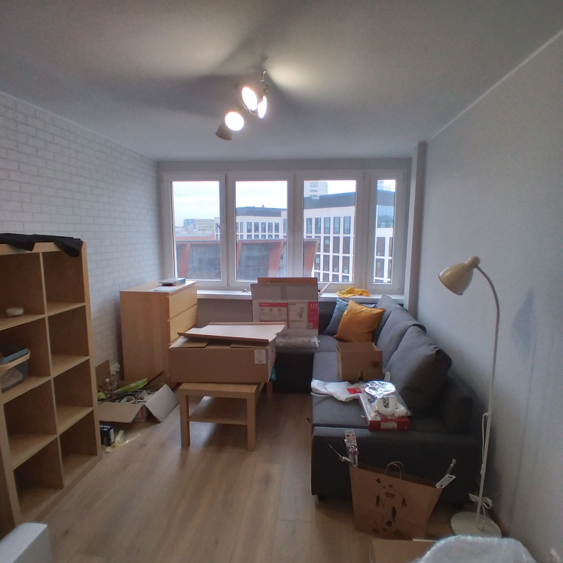 Pokój dzienny w trakcie przeprowadzki lub remontu, z kartonami, rozłożoną sofą i widocznymi elementami wykończenia podłogi.