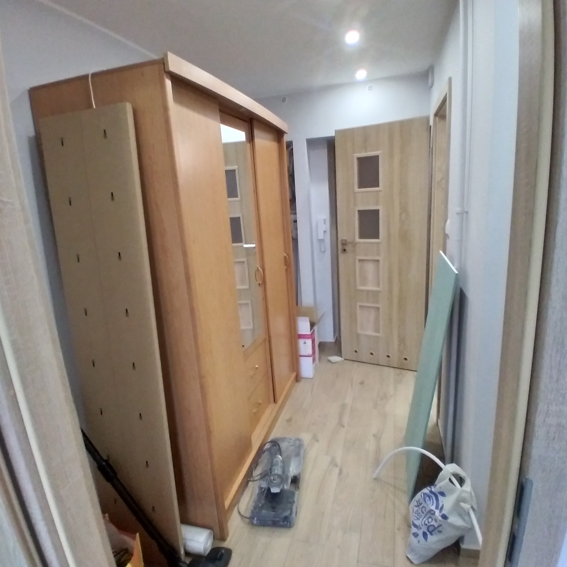 Montaż szafy przesuwnej w pokoju, widoczna płyta meblowa z otworami montażowymi, szlifierka kątowa na podłodze, przygotowania do montażu drzwi.