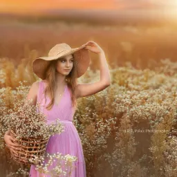 Młoda kobieta w zwiewnej sukience i kapeluszu trzyma koszyk pełen polnych kwiatów na tle łąki o zachodzie słońca.