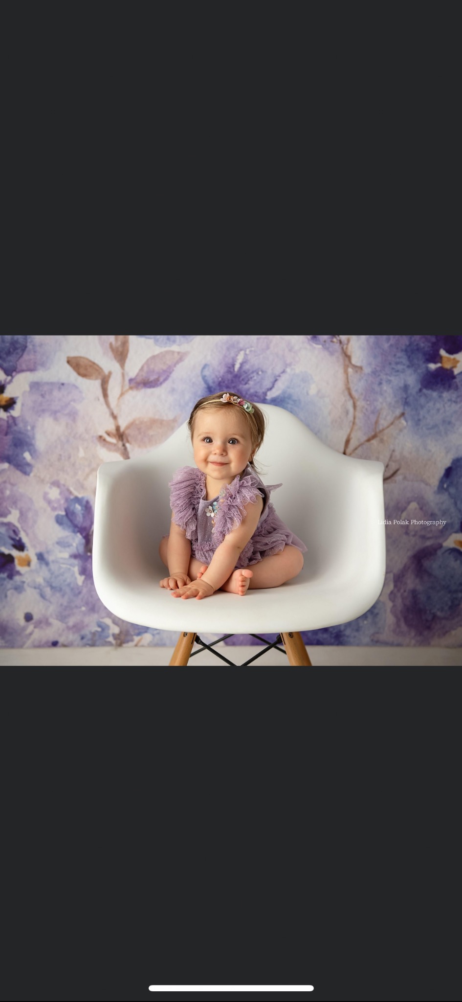 Uśmiechnięta dziewczynka w liliowym ubranku siedzi na białym krześle na tle fioletowych kwiatów.