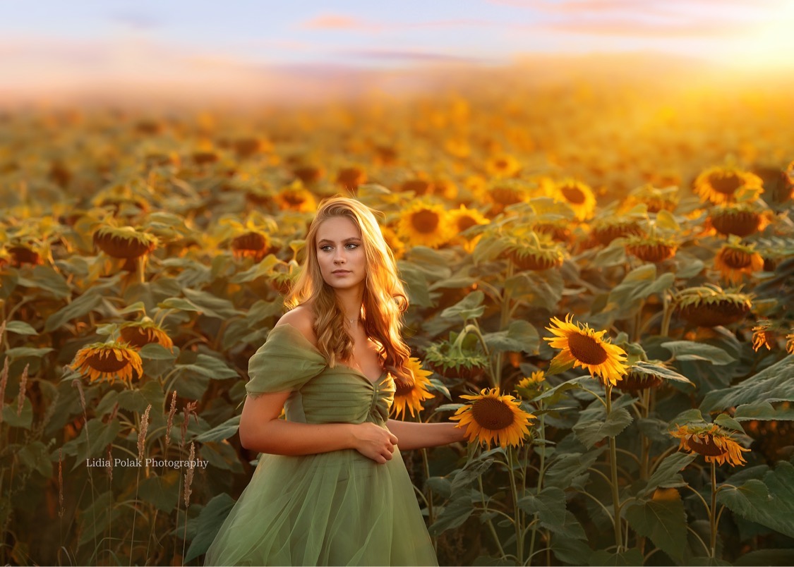 Młoda kobieta w zwiewnej, zielonej sukience pozuje na tle pola słoneczników o zachodzie słońca, delikatne światło podkreśla blond włosy.