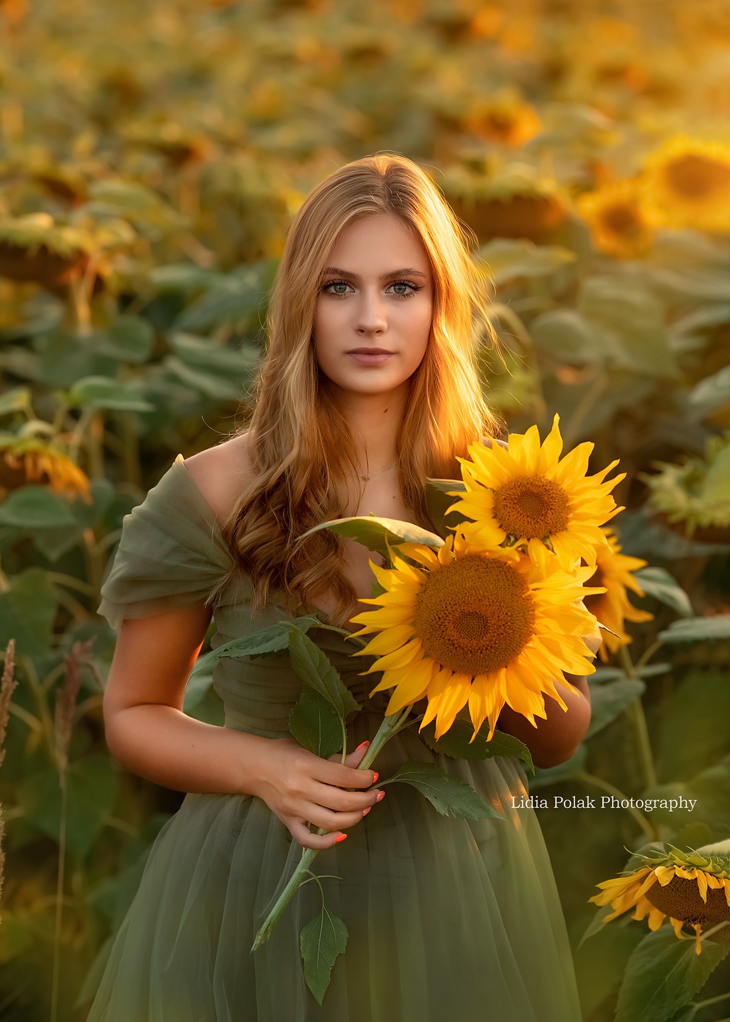 Portret młodej kobiety w zielonej sukience, trzymającej bukiet słoneczników na polu słoneczników w Jarosławiu, z pomalowanymi na pomarańczowo paznokciami.