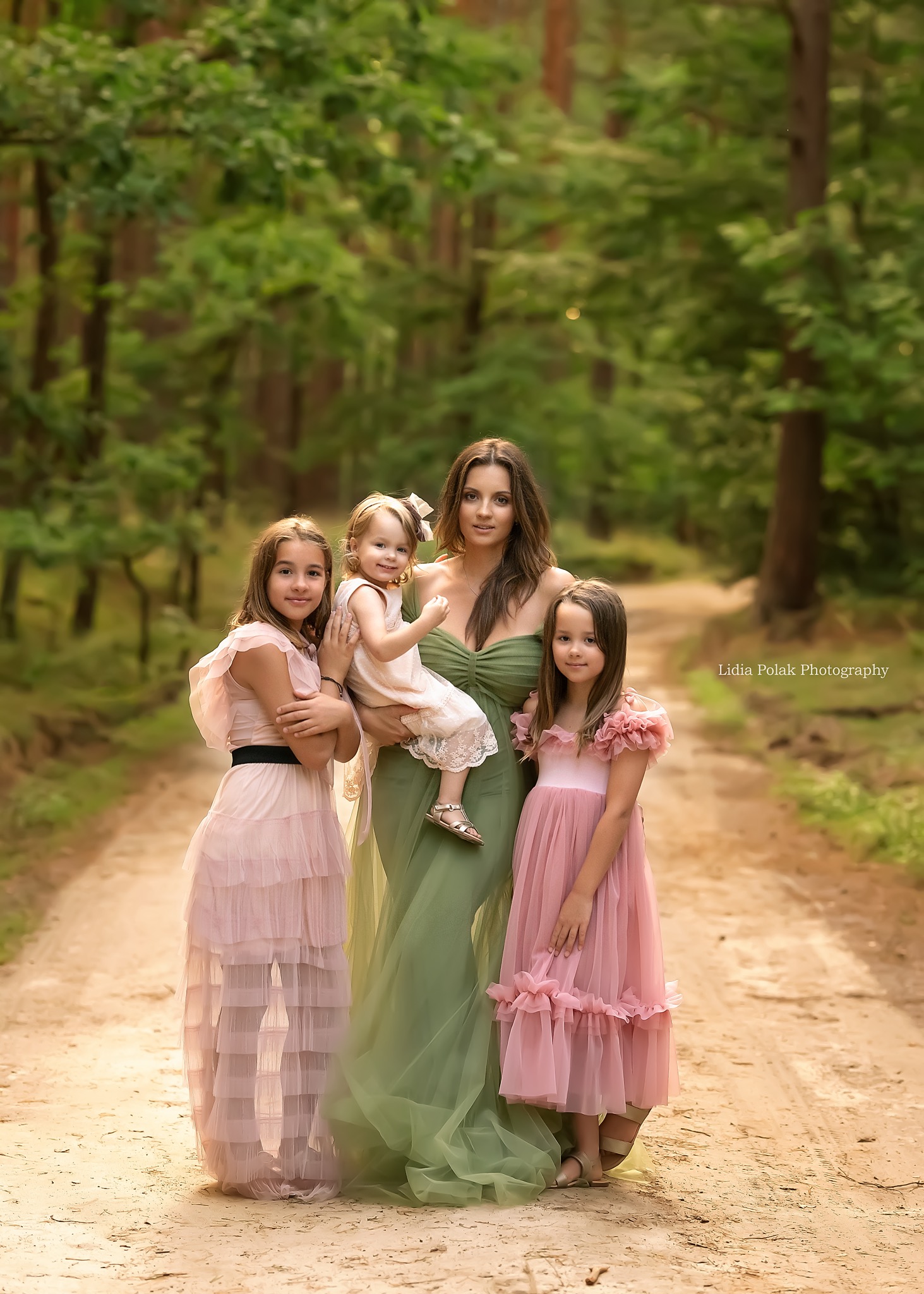Kobieta w długiej zielonej sukni trzymająca małą dziewczynkę, obok stoją dwie starsze dziewczynki w różowych sukienkach na leśnej ścieżce.