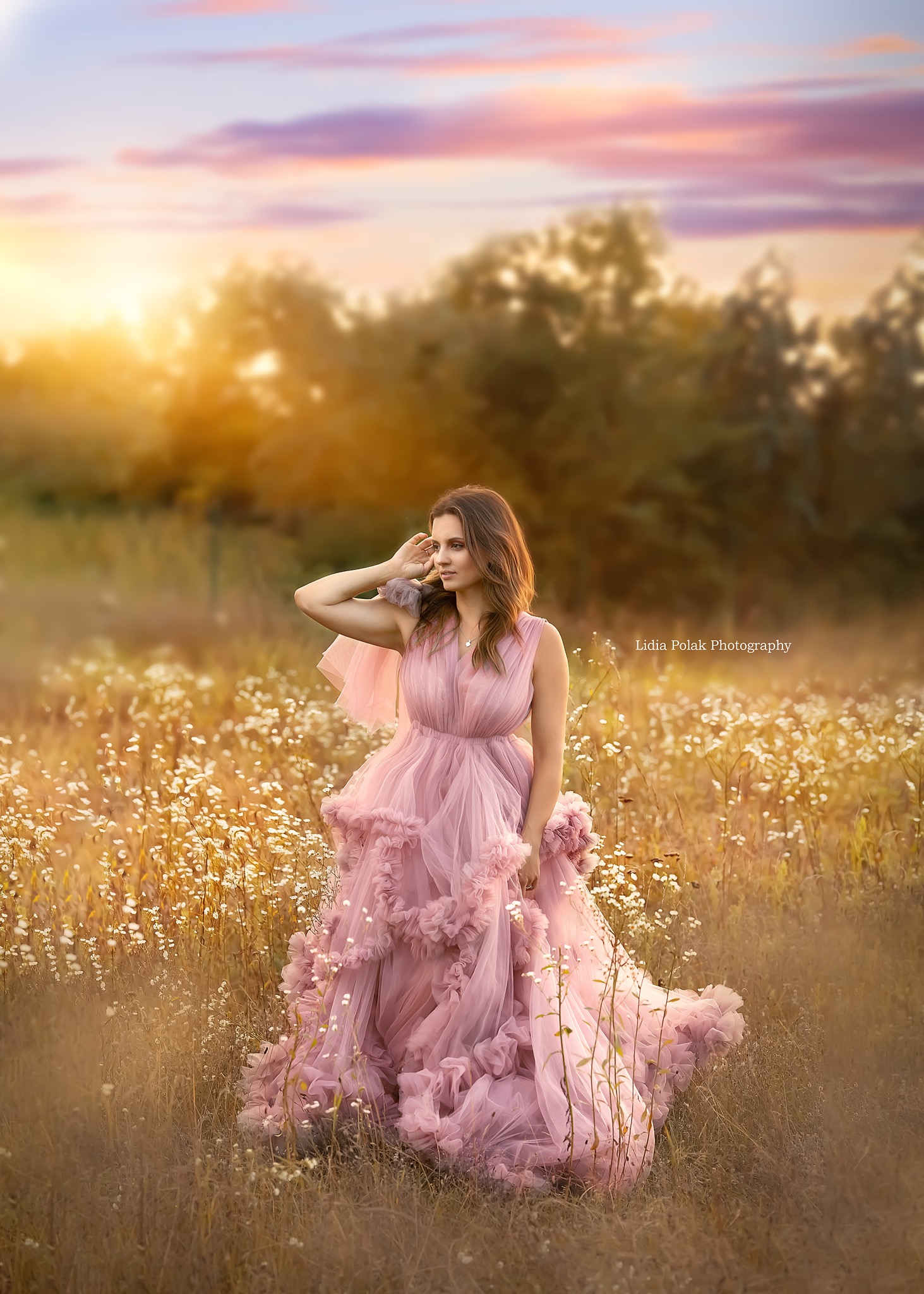 Kobieta w zwiewnej, różowej sukni z falbanami pozuje na łące pełnej białych kwiatów podczas zachodu słońca.