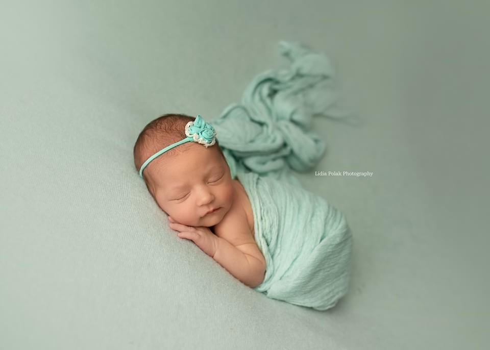 Śpiąca noworodka owinięta w miętowy kocyk z ozdobną opaską na głowie, leżąca na miętowym tle.