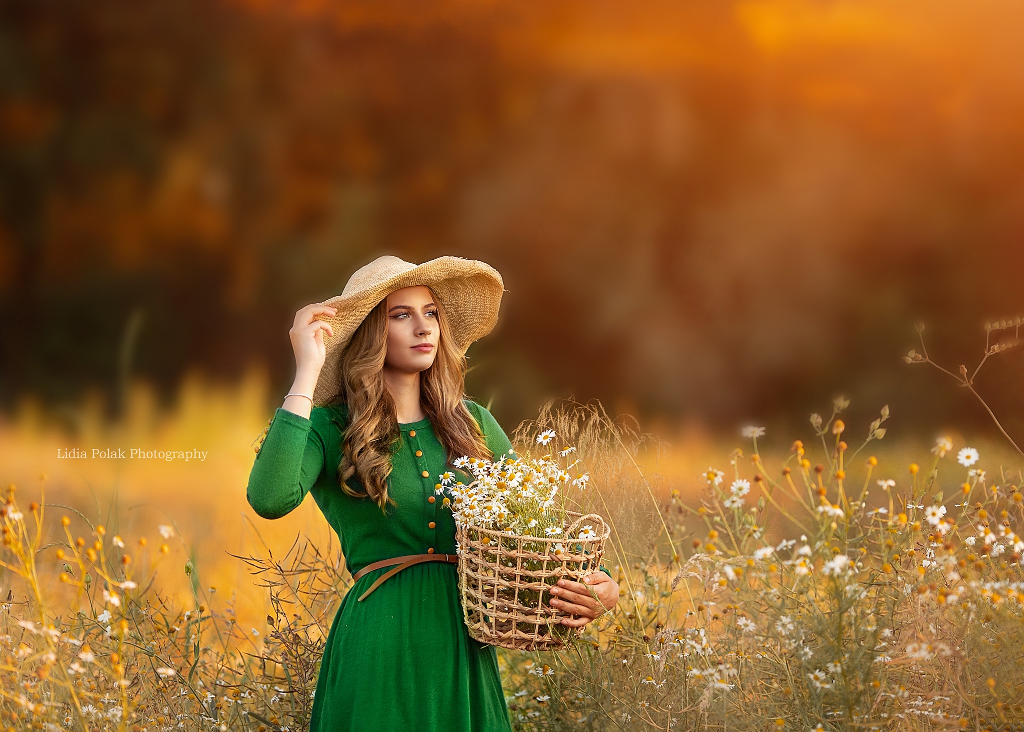 Portret kobiety w zielonej sukience i słomkowym kapeluszu, trzymającej koszyk z margaretkami na łące w ciepłym, złotym świetle.