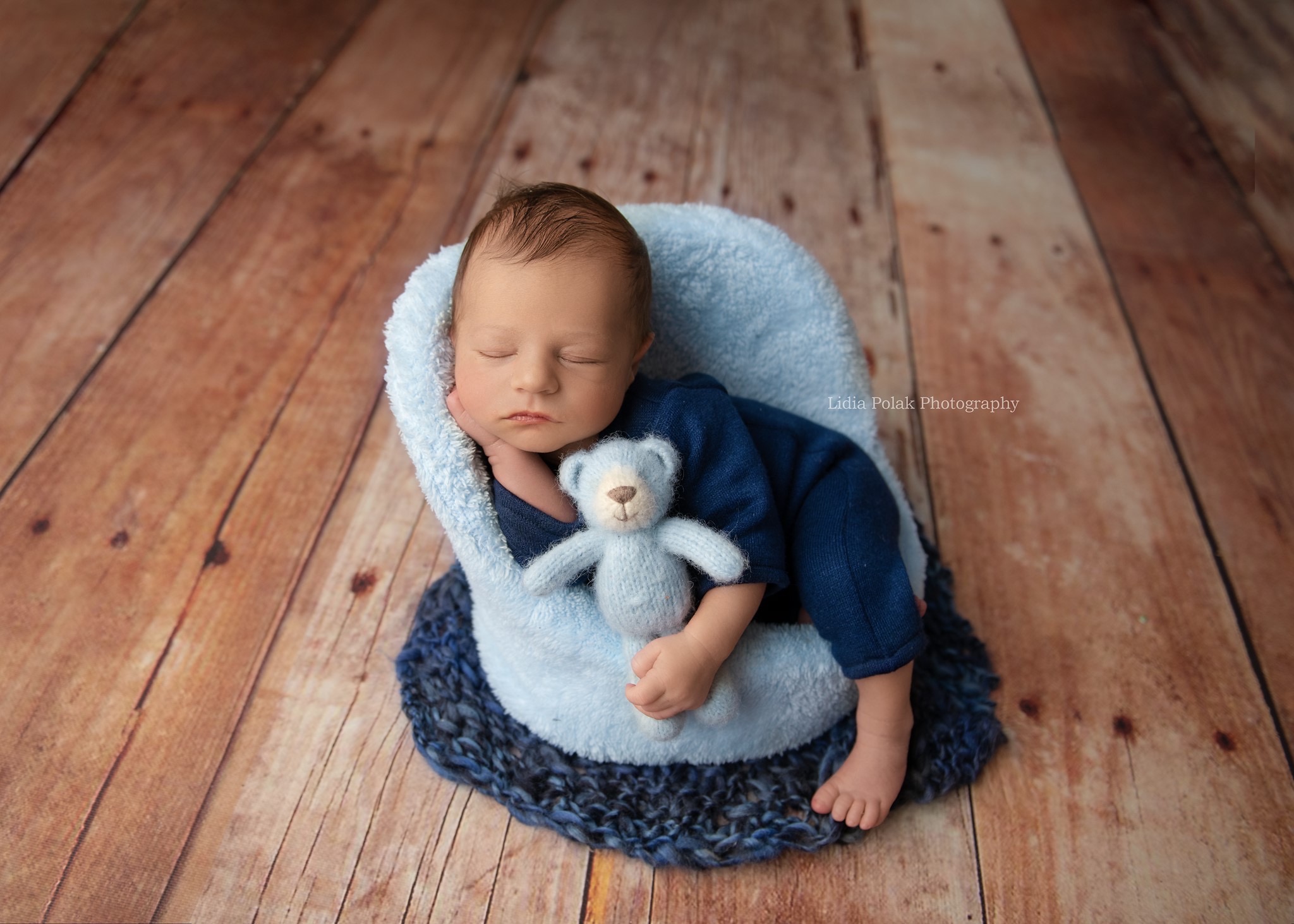 Śpiący noworodek w niebieskim ubranku, otulony miękkim, błękitnym kocykiem, trzymający pluszowego misia na drewnianym tle.