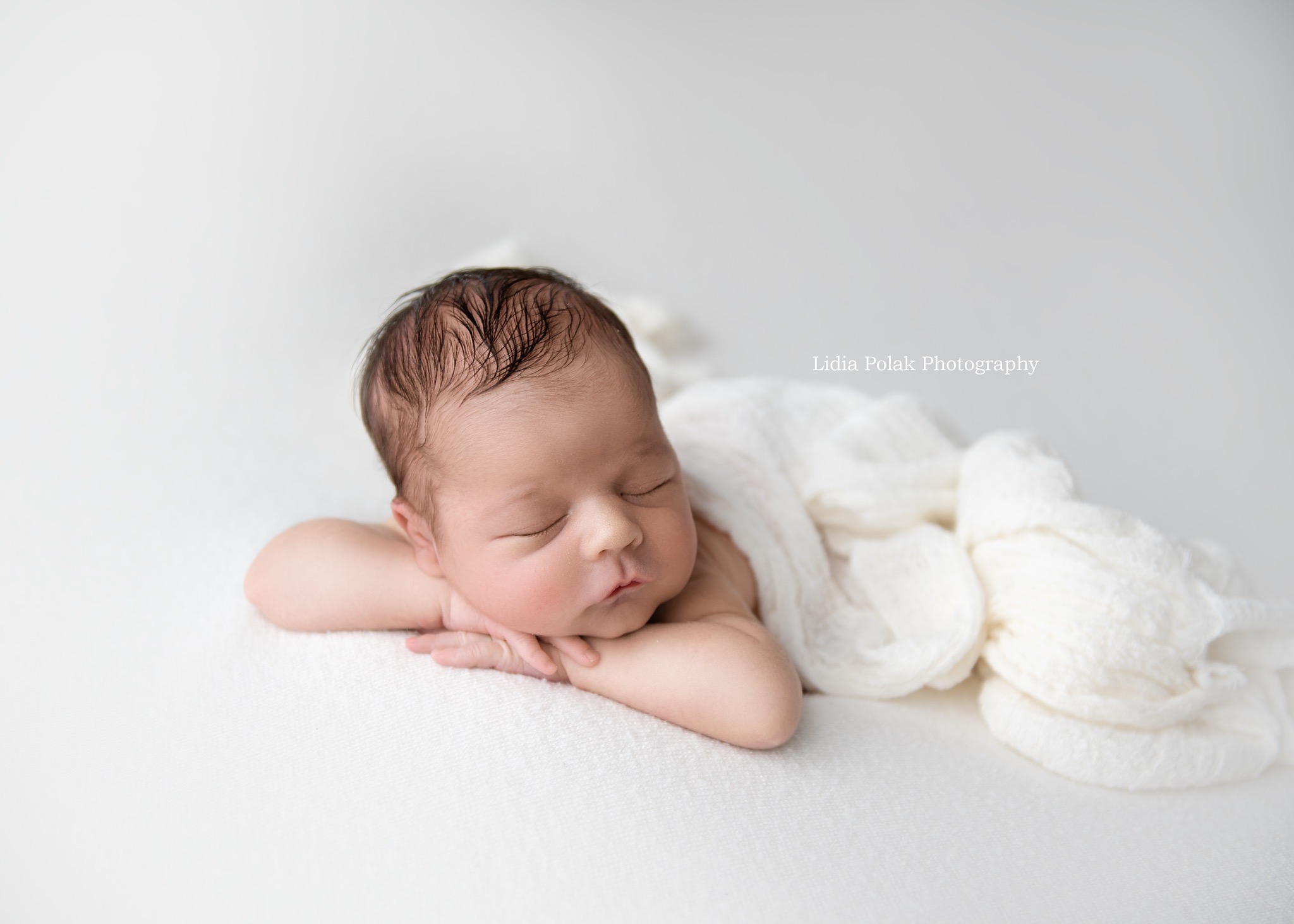 Noworodek śpiący na białym kocyku, z rączkami pod policzkiem, w delikatnym oświetleniu.