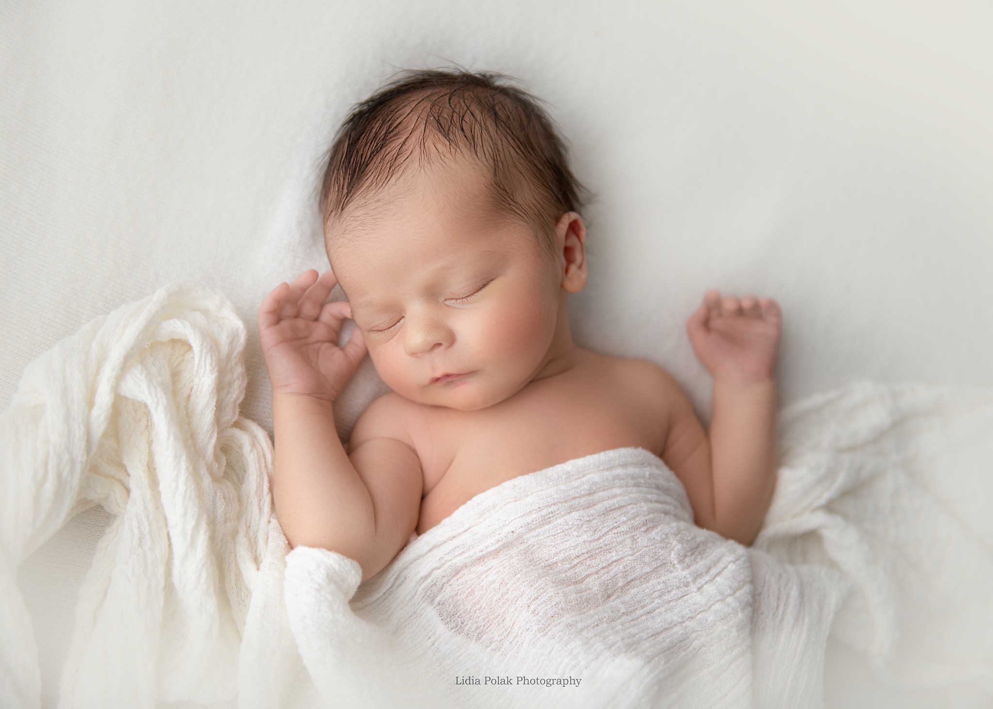 Śpiący noworodek zawinięty w białą, teksturowaną tkaninę, leżący na miękkim, białym tle, z delikatnym oświetleniem podkreślającym rysy twarzy i drobne dłonie.