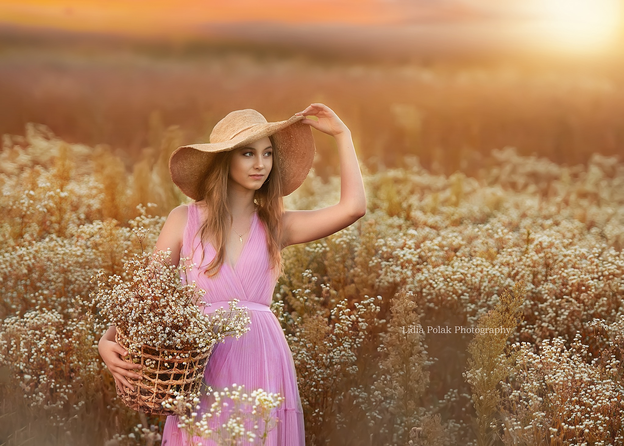 Młoda kobieta w zwiewnej sukience i kapeluszu trzyma koszyk pełen polnych kwiatów na tle łąki o zachodzie słońca.