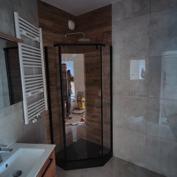 Marcin Jarosz - Nowoczesna łazienka z kabiną prysznicową z czarnymi profilami, umywalką z szafką z drewnopodobnym wykończeniem, grzejnikiem drabinkowym i szarymi płytkami na ścianach i podłodze. W lustrze kabiny...