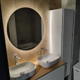 Nowoczesna łazienka z dwiema umywalkami nablatowymi, okrągłym lustrem z podświetleniem LED i dekoracyjną ścianą z wzorem jodełki. Minimalistyczny design i eleganckie wykończenie.