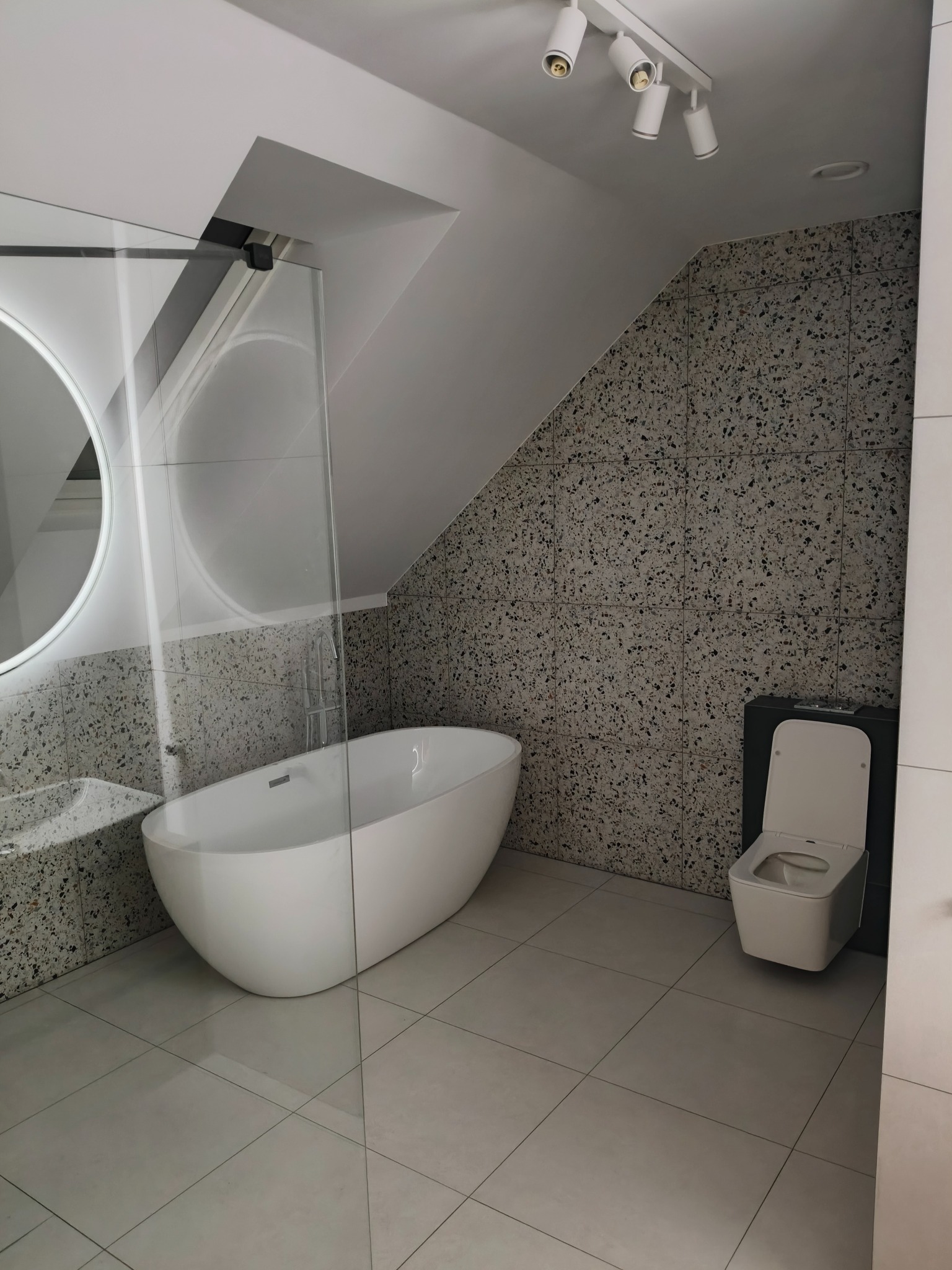 Nowoczesna łazienka na poddaszu z białą wolnostojącą wanną, kabiną prysznicową ze szklaną ścianką, okrągłym lustrem z podświetleniem LED i toaletą w zabudowie, wykończona płytkami imitującymi...