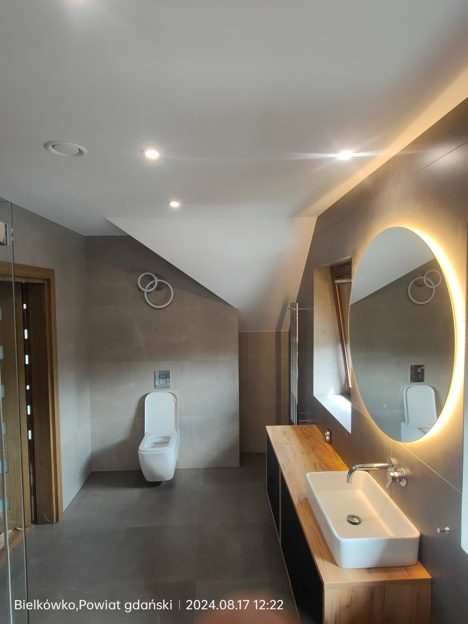 Nowoczesna łazienka na poddaszu z szarymi płytkami, wiszącą toaletą, umywalką nablatową na drewnianym blacie i okrągłym lustrem z podświetleniem LED.