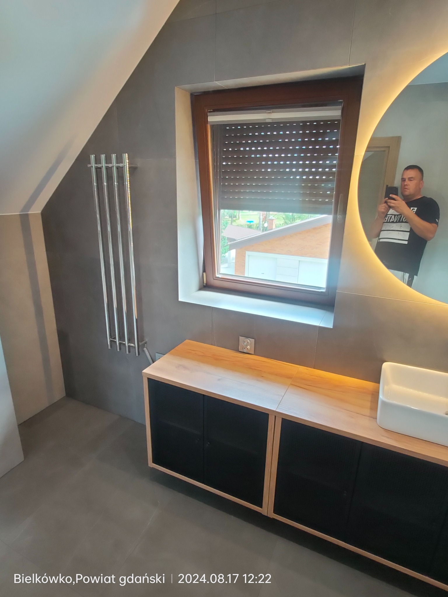 Nowoczesna łazienka na poddaszu z szarymi płytkami, grzejnikiem drabinkowym, szafką pod umywalkę z drewnianym blatem i okrągłym lustrem z podświetleniem LED, w którym odbija się osoba robiąca zdjęcie.