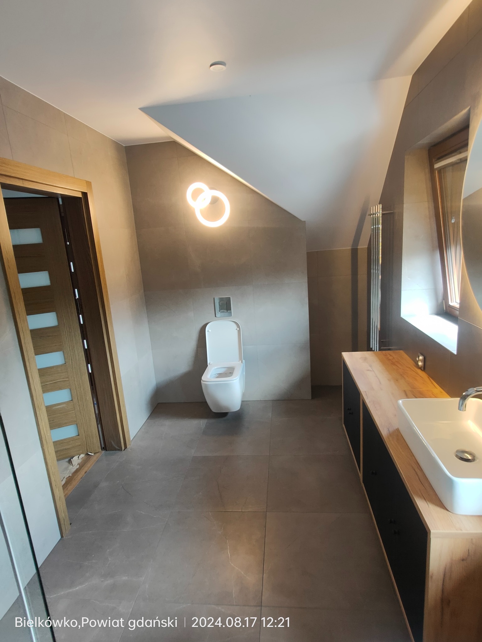 Nowoczesna łazienka na poddaszu ze skośnym sufitem, szare płytki podłogowe i ścienne, podwieszana toaleta, drewniana szafka z umywalką oraz okrągła lampa sufitowa.