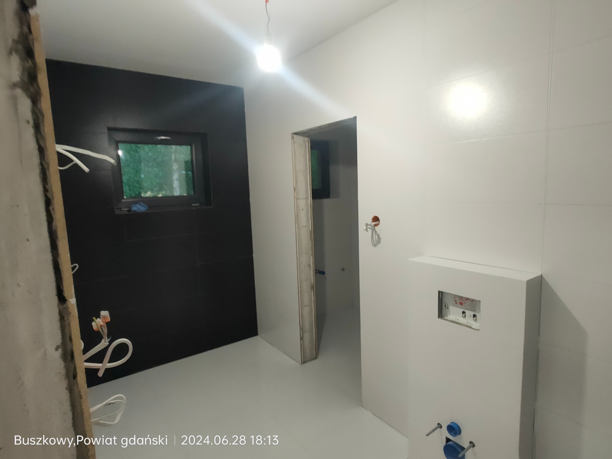Wnętrze łazienki w trakcie prac wykończeniowych: czarna ściana z oknem, białe płytki, podtynkowa instalacja sanitarna, widoczne przewody elektryczne i surowy fragment ściany.