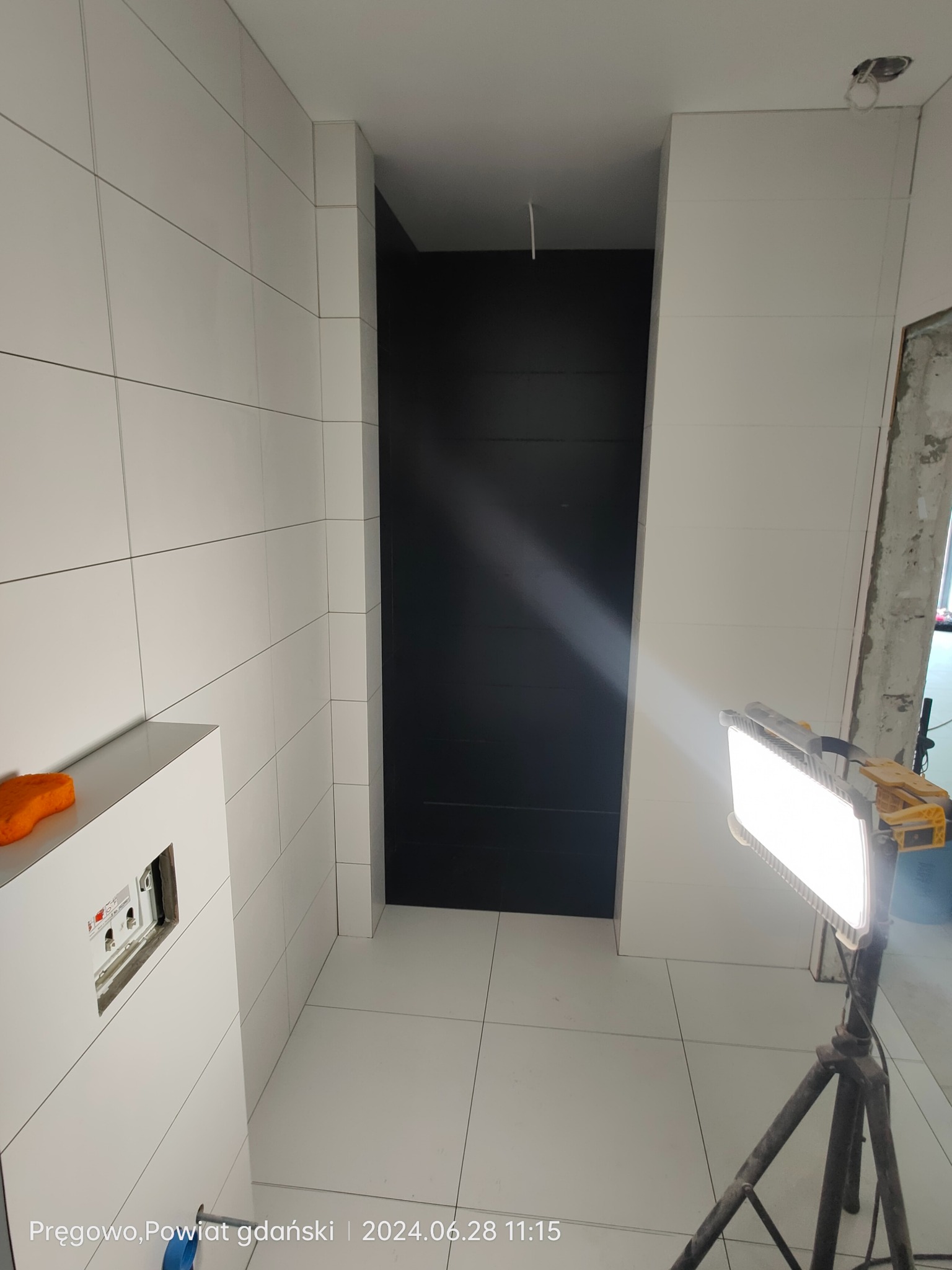 Wnętrze łazienki w trakcie prac wykończeniowych, widoczne białe płytki na ścianach i podłodze, czarne wnęki, instalacja podtynkowa WC oraz lampa budowlana na statywie.