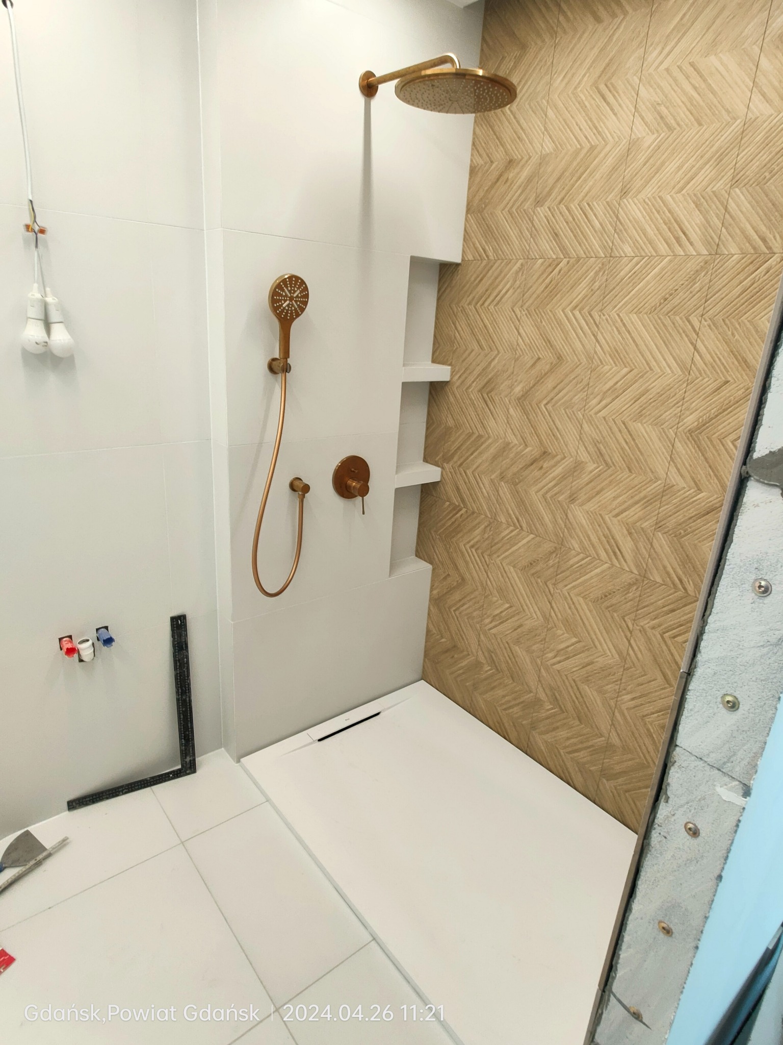 Łazienka w trakcie remontu: widoczna strefa prysznicowa z białą brodzikiem, ścianą z białych płytek i ścianą wyłożoną płytkami imitującymi drewno ułożonymi we wzór jodełki, a także instalacja...