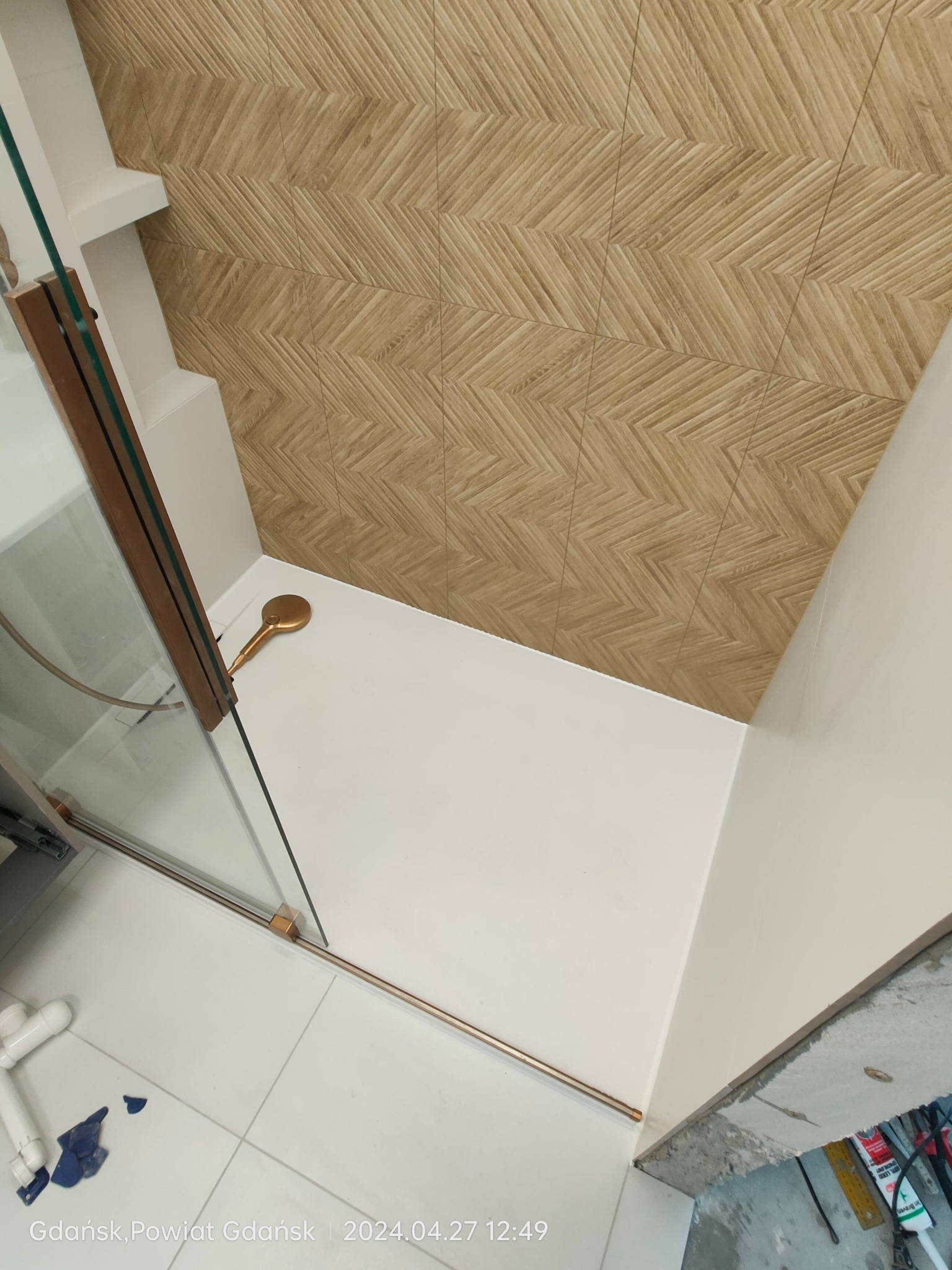 Kabina prysznicowa z drewnopodobnymi płytkami na ścianie, widoczna złota słuchawka prysznicowa i biała podłoga z fugami, dolny kadr z widocznymi elementami instalacji przed wykończeniem.