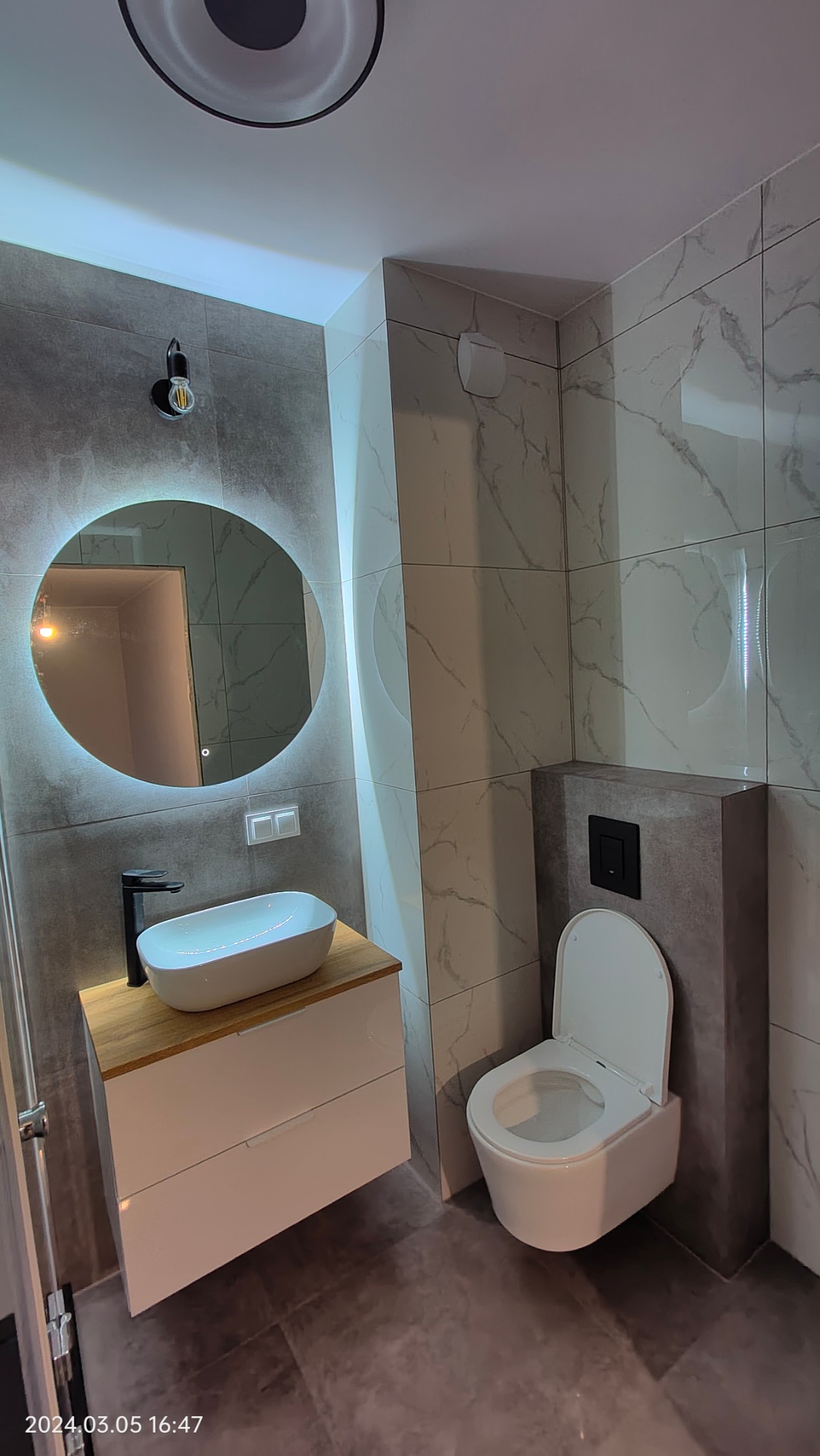 Nowoczesna łazienka z szarymi płytkami imitującymi marmur, okrągłym lustrem z podświetleniem LED, wiszącą szafką z umywalką nablatową i czarną armaturą oraz podwieszaną toaletą.