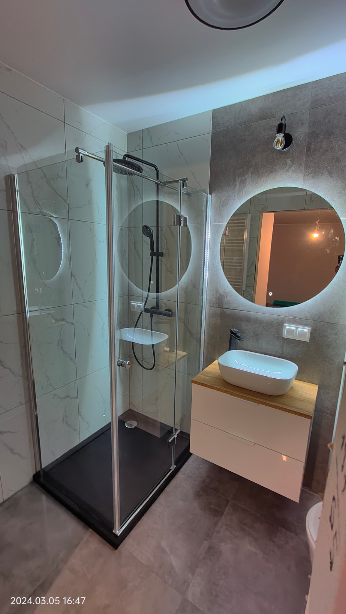 Nowoczesna łazienka z kabiną prysznicową typu walk-in z czarnym brodzikiem, okrągłym lustrem z podświetleniem LED i umywalką nablatową na szafce z drewnianym blatem.