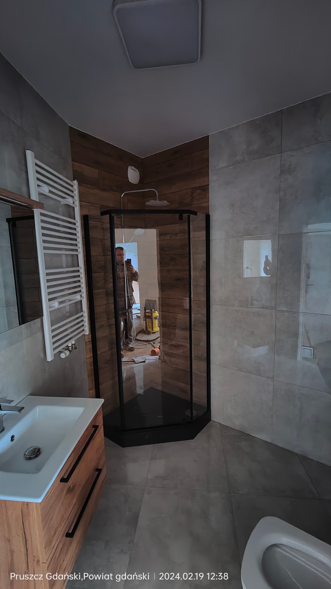 Nowoczesna łazienka z kabiną prysznicową z czarnymi profilami, umywalką z szafką z drewnopodobnym wykończeniem, grzejnikiem drabinkowym i szarymi płytkami na ścianach i podłodze. W lustrze kabiny...