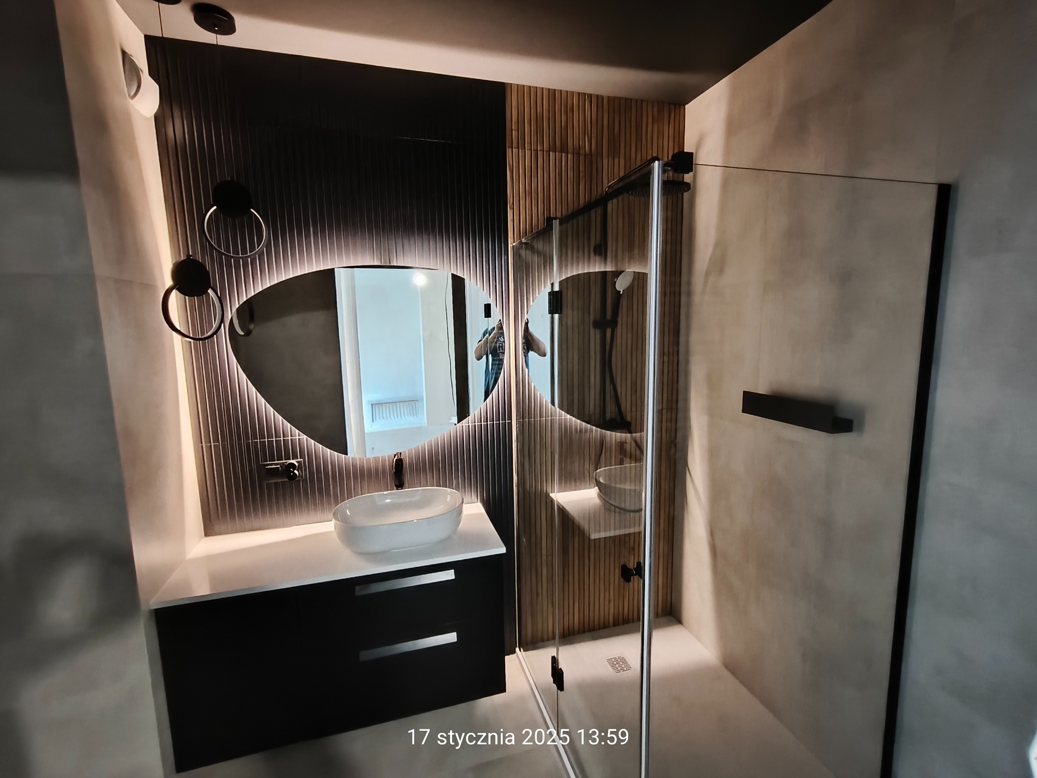 Nowoczesna łazienka z podświetlanym lustrem o owalnym kształcie, czarną szafką pod umywalką i kabiną prysznicową ze szklanymi ścianami, wykończona płytkami w odcieniach szarości i drewna.