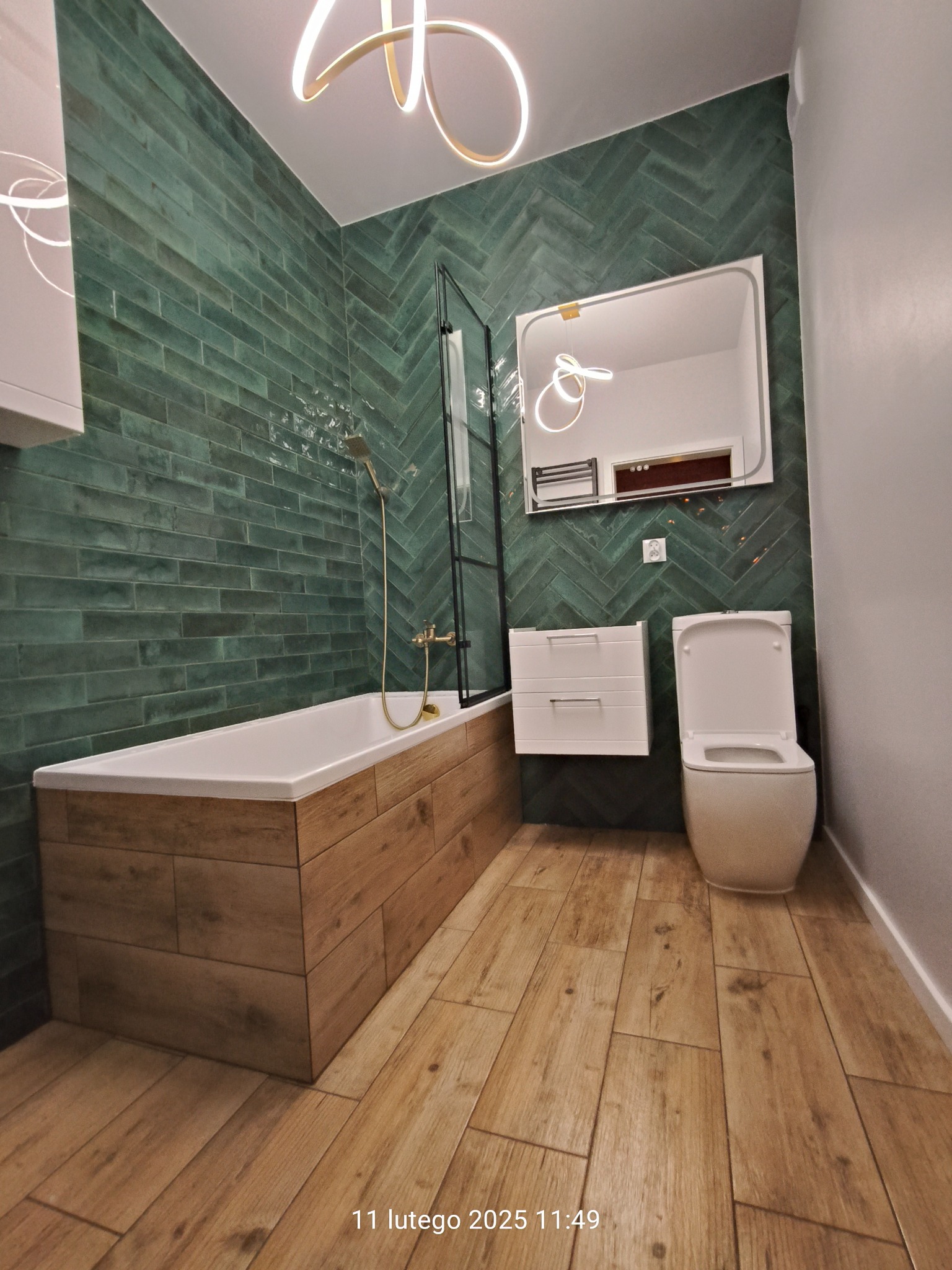 Nowoczesna łazienka z zielonymi płytkami ułożonymi w jodełkę, obudową wanny imitującą drewno, wiszącą szafką z umywalką i podświetlanym lustrem. Na podłodze płytki imitujące drewno.