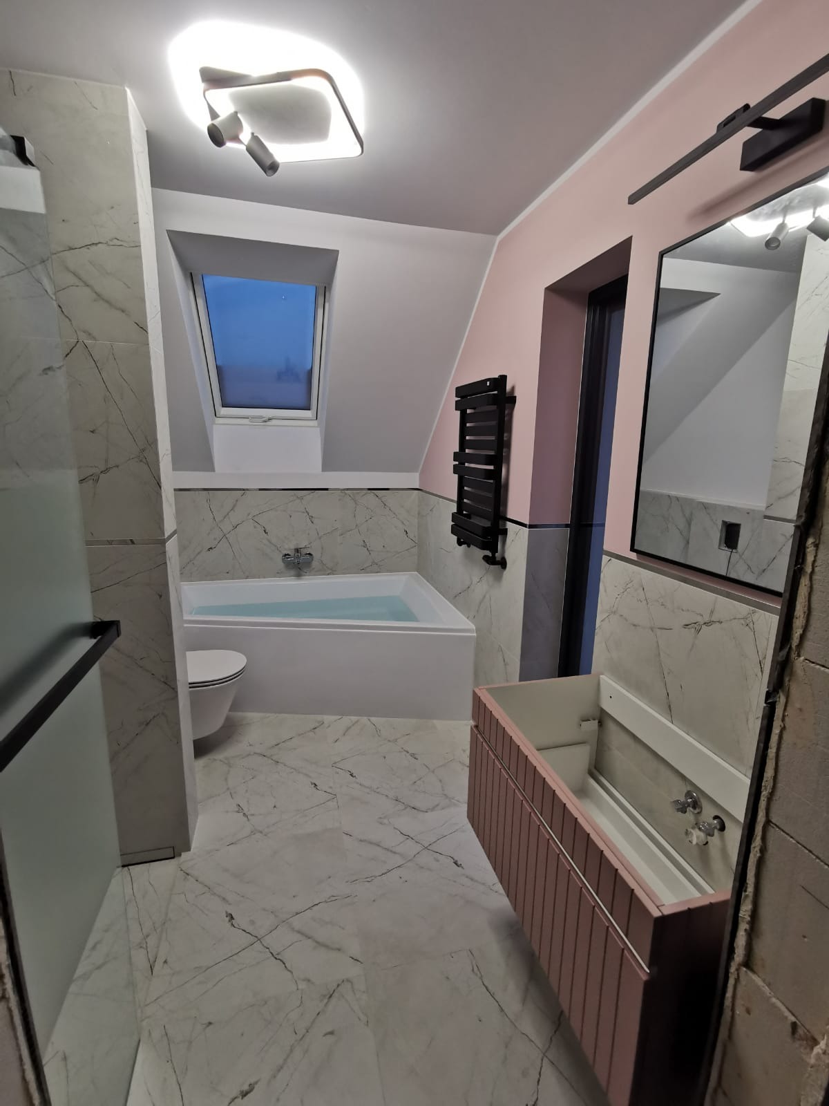 Nowoczesna łazienka na poddaszu z marmurowymi płytkami, wanną wypełnioną wodą i oknem dachowym. Różowe akcenty kolorystyczne na ścianie i szafce pod umywalką.