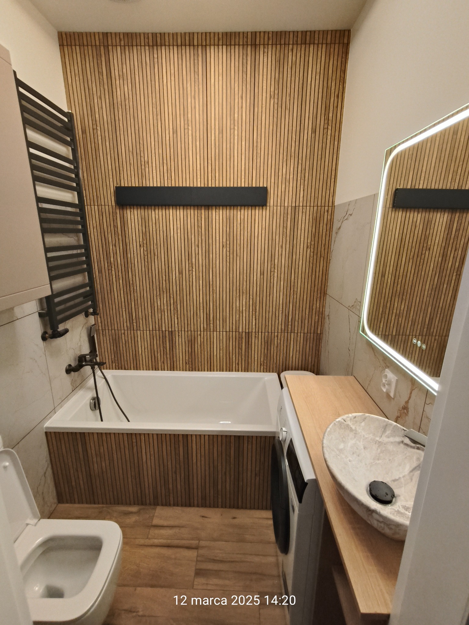 Nowoczesna łazienka po remoncie: drewniane lamele na ścianie, podświetlane lustro, marmurowa umywalka, pralka i grzejnik drabinkowy. Wykończenie w ciepłych barwach.