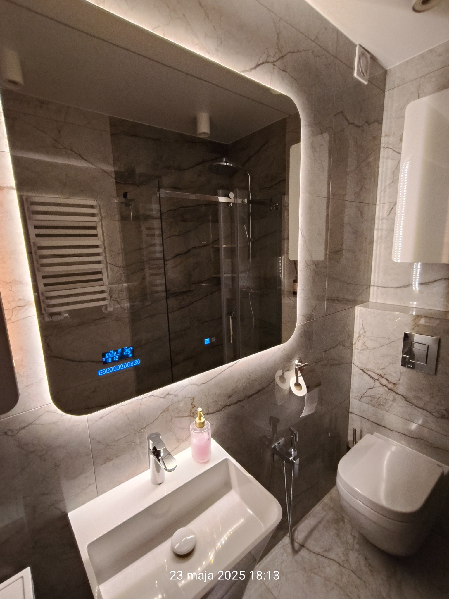 Wykończona łazienka w szarościach. Umywalka z baterią, toaleta, bidetka i podświetlane lustro z wyświetlaczem. Kabina prysznicowa widoczna w odbiciu lustra.