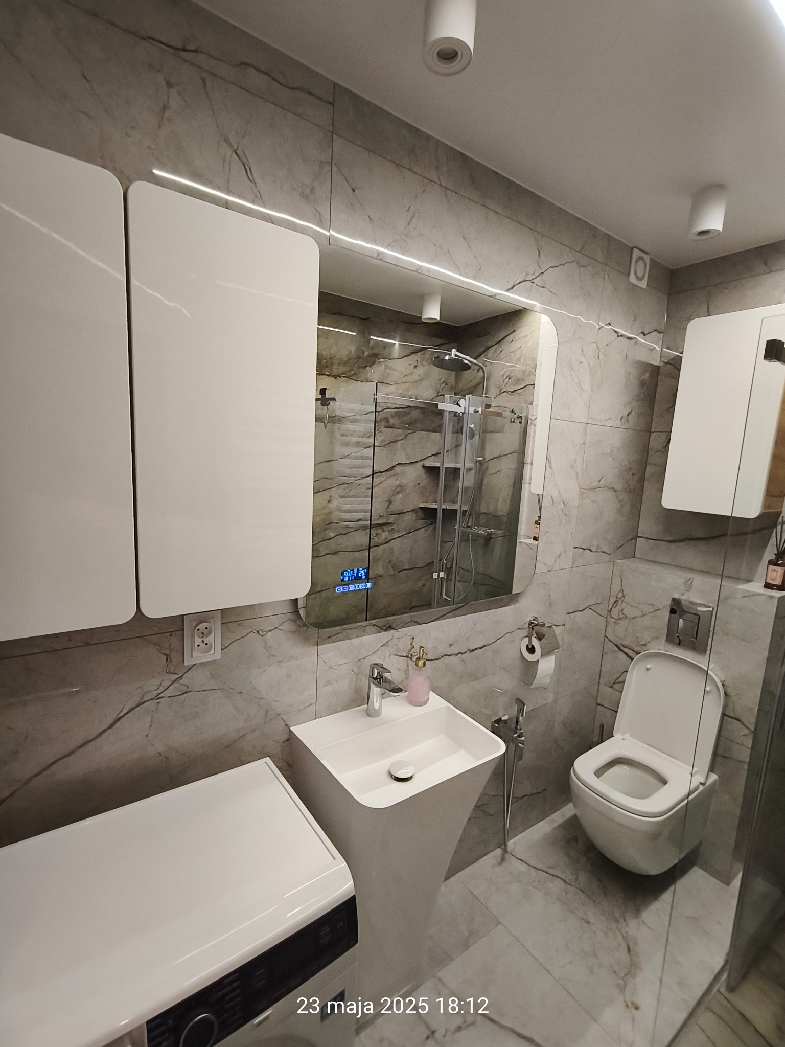 Nowoczesna łazienka w szarościach z podświetlanym lustrem, białą umywalką na postumencie i wiszącą toaletą. Marmurowe płytki na ścianach i podłodze.