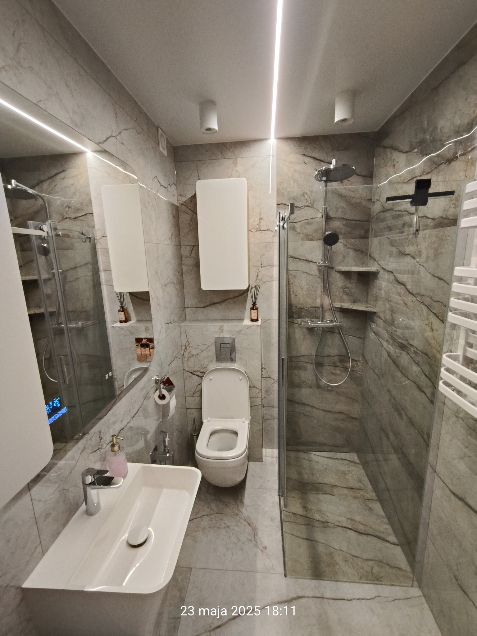 Nowoczesna łazienka z marmurowymi płytkami, kabiną prysznicową typu walk-in, podświetlanym lustrem i wiszącą szafką. Minimalistyczny design z akcentami zapachowymi.