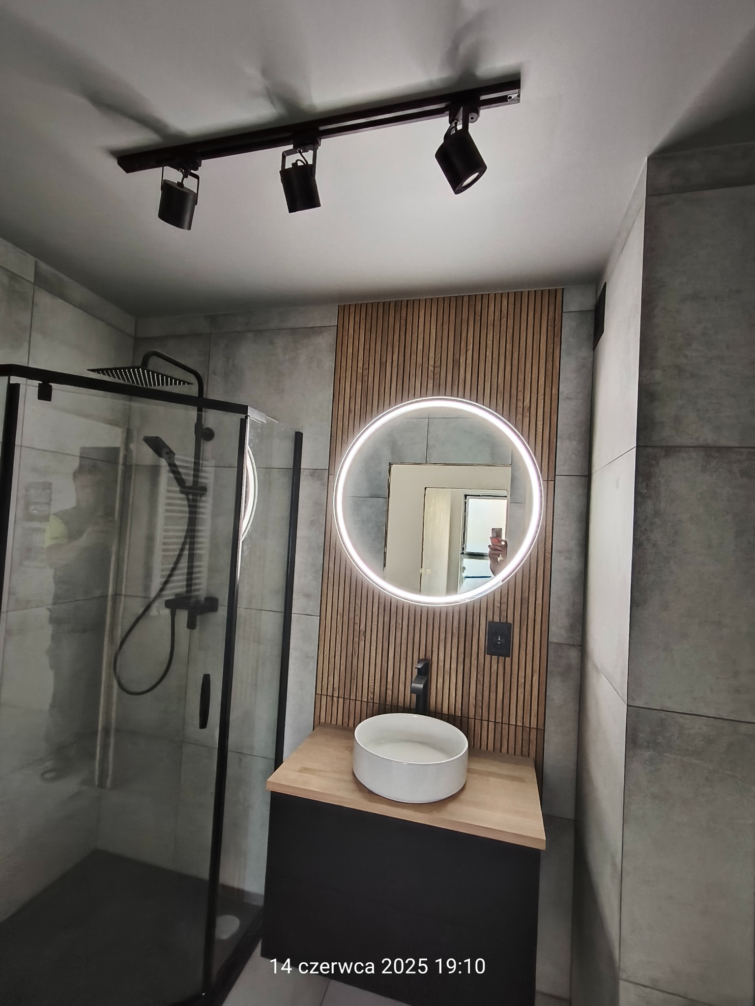 Nowoczesna łazienka w Pręgowie Dolnym: szara glazura, kabina prysznicowa z czarnym wykończeniem, okrągłe lustro LED i drewniana lamela ścienna.