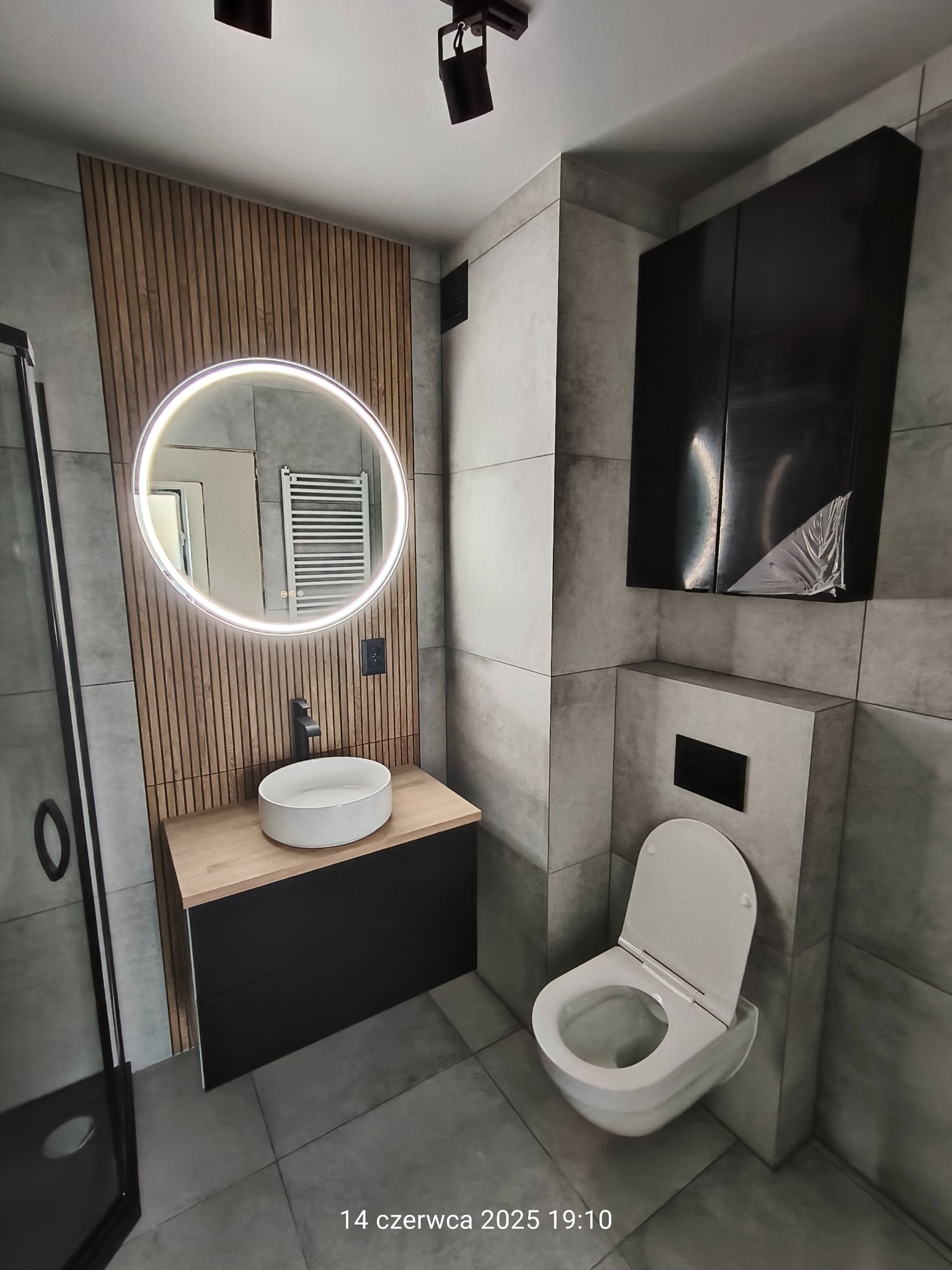 Nowoczesna łazienka w szarościach z drewnianymi akcentami: okrągłe lustro z podświetleniem, wisząca szafka, toaleta i kabina prysznicowa.